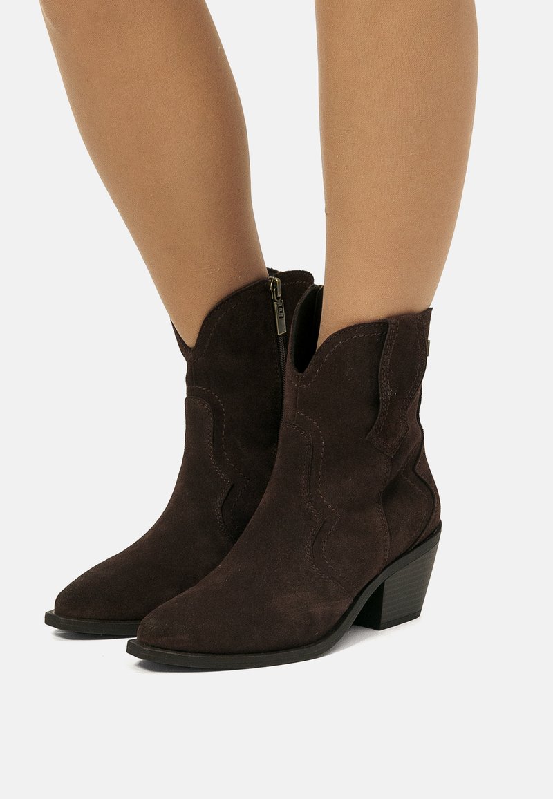 Stivali alla caviglia in suede marrone scuro con tacco basso impilato e cerniere laterali indossati su gambe nude contro uno sfondo semplice.