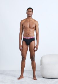 Černé bavlněné slipy s červenobílým logem Nike na elastickém pásku, zobrazené na mužském modelu stojícím na světle šedém povrchu před bílým pozadím.