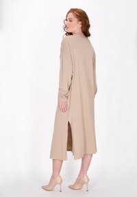 Beige, hosszú ujjú, A-vonalú ruha oldalsó hasítékokkal, laza szabással és sima textúrával. A modellen meztelen színű magassarkú cipő van.