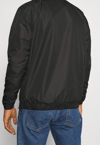 Veste légère noire avec poignets et ourlet élastiques, présentant une texture lisse et mate. Dotée d'un design minimaliste, sans marques visibles.