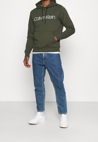 Felpa con cappuccio verde oliva con logo "Calvin Klein" bianco, indossata sopra una camicia bianca. Abbinata a jeans blu e sneakers bianche.