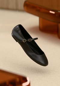 Schwarzer Ballerina-Schuh aus Leder mit abgerundeter Spitze und einem dünnen Knöchelriemen mit goldfarbenem, rundem Schnallenakzent. Glatte Textur und minimalistisches Design.