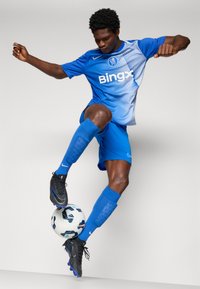 Blue Nike football kit with geometric pattern, matching shorts, and blue socks. Black cleats with blue accents; player controls a patterned soccer ball.