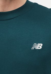 Mann trägt ein dunkelgrünes T-Shirt mit Rundhalsausschnitt und einem kleinen weißen New Balance-Logo auf der Brust.