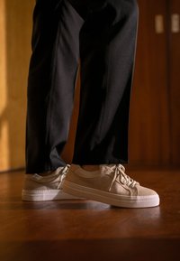 Sneakers beige in suede con finitura texturizzata, punta rotonda e suola in gomma bianca. I lacci sono di un taupe chiaro, abbinati a pantaloni neri. Sfondo in parquet.