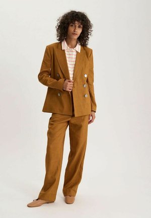 Femme aux cheveux bouclés debout, portant un costume croisé moutarde, une chemise rayée col et des chaussures plates beiges sur un fond uni.