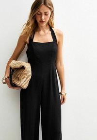 Mono negro con escote cuadrado y tirantes de halter. Textura suave, piernas anchas. Se sostiene un bolso de mano de paja trenzada. Pulsera de oro en la muñeca.