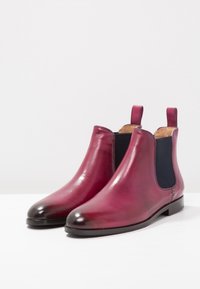 Pár lesklých tmavě červených kožených Chelsea bot s černými elastickými bočními panely a poutky na bílé ploše.