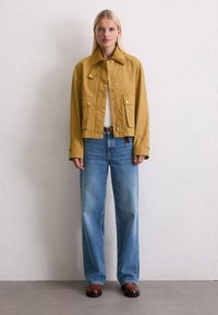 Khaki gekleurde jas met een klassieke kraag en drukknoopsluiting, in combinatie met lichtblauwe wijde jeans en bruine schoenen. Geweven stofdetail.