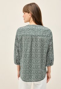 Grün gemusterte Bluse mit Dreiviertelärmeln, die ein geometrisches Design und einen gefältelten Rücken für zusätzliche Textur bietet.