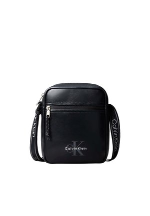 Mała czarna torba Calvin Klein na ramię z przednią kieszenią na zamek i regulowanym paskiem na ramię z logo na białym tle.