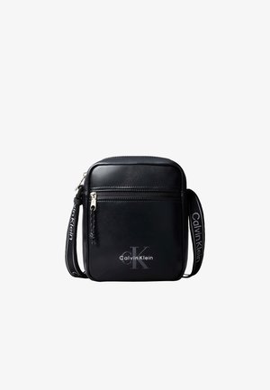 Petit sac bandoulière noir Calvin Klein avec poche zippée à l'avant et bandoulière réglable de marque sur fond blanc.