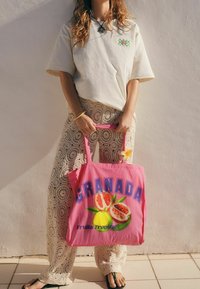 Sac fourre-tout rose avec le texte "GRANADA" en grandes lettres bleues, accompagné d'illustrations de fruits. Mannequin portant un haut beige et un pantalon à motif dentelle.