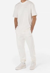 Hombre vistiendo un conjunto casual completamente blanco con una camisa de manga corta, pantalones jogger y zapatillas blancas, con las manos en los bolsillos, de pie.
