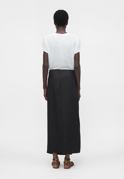 Tagant vaadatud inimene, kes kannab valget lühikeste varrukatega särk, pikka musta seelikut ja pruune pahlaga paeltega sandaale, seisab ühtlasel taustal.