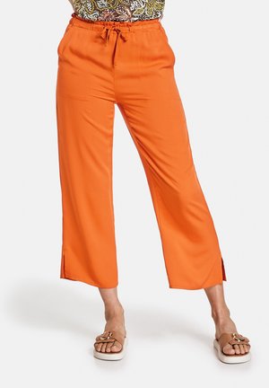Femme portant un pantalon large court orange vif avec des fentes latérales et des sandales marron, debout les mains sur les hanches.