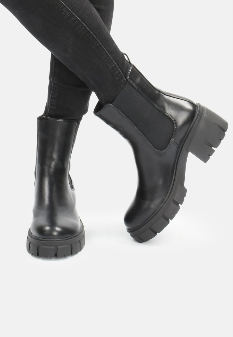 Botas negras hasta el tobillo hechas de material brillante, con suela gruesa, paneles laterales elásticos y diseño de punta redondeada.