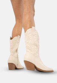 Kremowe skórzane kowbojki z dekoracyjnym haftem, spiczastym noskiem i lekko ukośnym, drewnianym obcasem.
