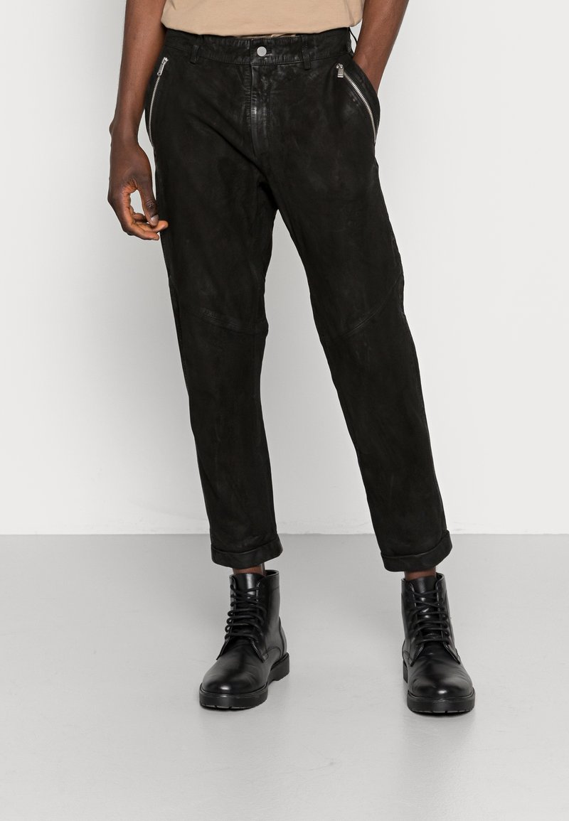 Homme portant un pantalon en daim noir avec poches zippées et revers retroussés, associé à des bottes en cuir noires à lacets sur un sol gris.