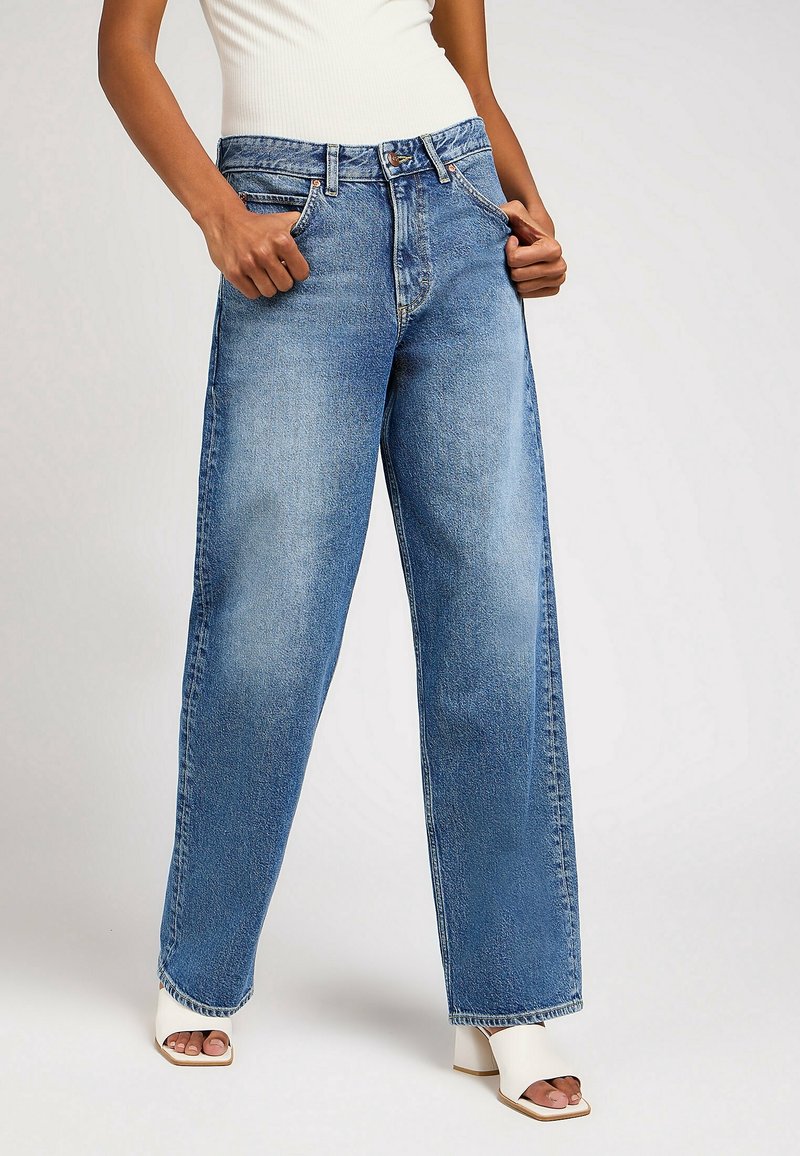 Jean bleu clair taille haute avec une coupe ample, arborant un style classique à cinq poches et un léger effet froissé, associé à des sandales blanches à talons carrés.