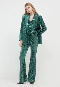 Completo di velluto verde smeraldo con giacca fitted e pantaloni a zampa, caratterizzato da una consistenza morbida e una leggera lucentezza, abbinato a décolleté nere con punta.