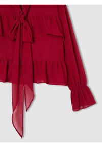 Blusa leggera rossa con nodo frontale, strati arricciati e maniche lunghe con polsini elasticizzati. Texture liscia e dettagli di sovrapposizione trasparente.