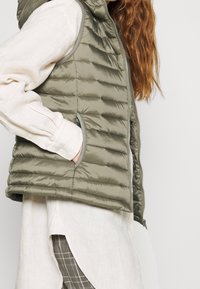 Khaki péřová vesta s prošívanou strukturou, bočními kapsami a zipovým zapínáním, nošená přes svetřík dlouhého rukávu v světlé barvě.