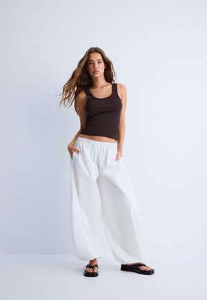Giovane donna in piedi con le mani in tasca, indossa un top nero senza maniche, pantaloni bianchi larghi e sandali neri con plateau su uno sfondo chiaro uniforme.
