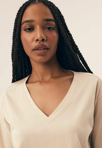 Beżowa koszulka z dekoltem w serek, wykonana z gładkiego materiału, z luźnym krojem i prostym wzorem. Modelka ma długie, zaplecione włosy.