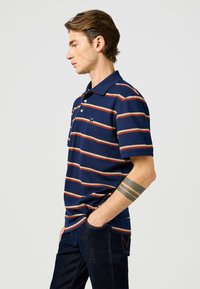 Marinblå randig piquétröja med horisontella orange och beige ränder, korta ärmar, en tre-knapps knäppning och en liten logotyp på bröstet.