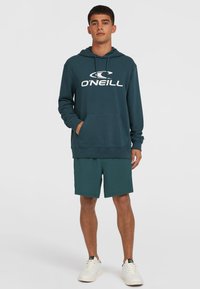 Tifana kapuca v teal barvi z velikim belim logotipom "O'Neill", z sprednjim žepom in kapuco s vrvico, v kombinaciji z usklajenimi šortami in belimi športnimi copati.