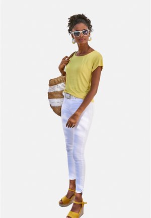 Gelbes Kurzarmoberteil, weiße Skinny-Jeans und gelbe Keil-Sandalen. Die Accessoires umfassen große weiße Sonnenbrillen und eine geflochtene Tragetasche.