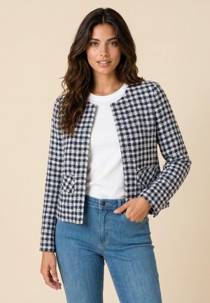 Donna con lunghi capelli scuri che indossa una camicia bianca, una giacca a quadri blu e bianca e jeans blu su uno sfondo beige.
