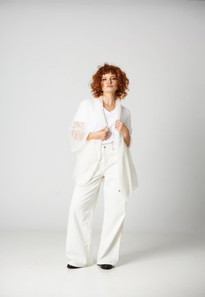 Ruda osoba stoi na jednolitym tle, ubrana w białe spodnie o szerokich nogawkach, biały top i luźny, teksturowany biały kardigan.