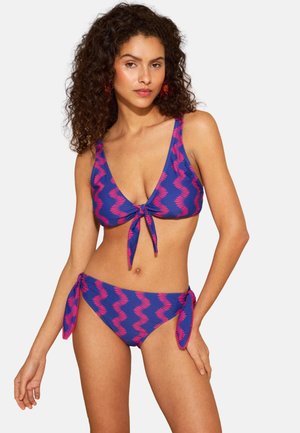 Kvinde med krøllet hår iført en lilla bikini med pink bølgede striber og bindedetaljer på top og bund står mod en ensfarvet baggrund.