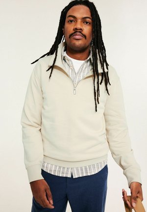 Mann mit langen Dreadlocks, der einen cremefarbenen Sweatshirt mit Viertelreißverschluss über einem gestreiften Hemd und marineblauen Hosen trägt und ein beigefarbenes Objekt hält.