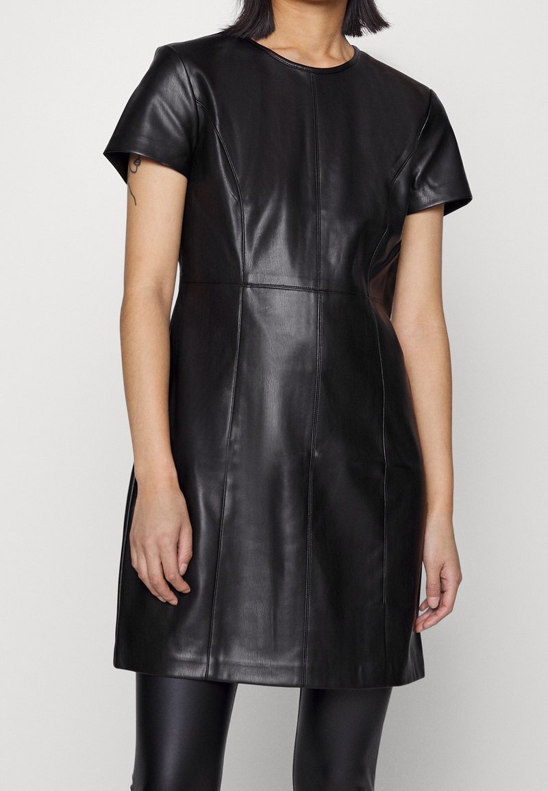 Femme portant une robe en cuir noire à manches courtes avec des détails de couture et des leggings noirs, vue des épaules jusqu'à la mi-cuisse.