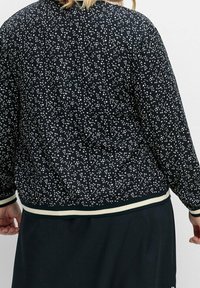 Giacca bomber nera con un motivo a pois bianchi, polsini e orlo a coste color crema. Tessuto morbido con una vestibilità comoda.