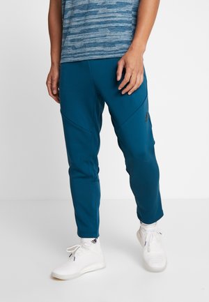 Homme portant un pantalon de sport bleu et des baskets blanches, debout devant un fond blanc uni.