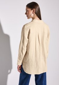 Béžová košile s dlouhým rukávem vyrobená z texturované látky, s límečkem na knoflíky a skladem na zadní straně, doplněná tmavě modrými džíny.