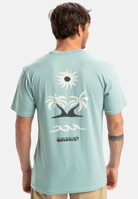 Světle modré tričko s grafickým designem slunce, palem a vln v bílé a černé barvě. Název značky "Quiksilver" je součástí designu.