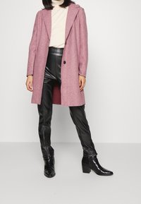 Nainen, jolla on päällään vaaleanpunainen villakangastakki, beige poolopaita, mustat nahkahousut ja mustat korkokengät nilkassa, seisoo valkoista taustaa vasten.