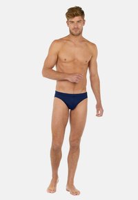 Námořnické modré plavky z pružného materiálu, s bezešvou gumou v pase a minimálním krytím, nosí je mužský model.