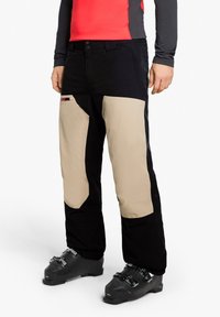 Black and beige ski pants with reinforced knee patches, featuring a front button closure and side pocket detail. Suitable for winter sports.