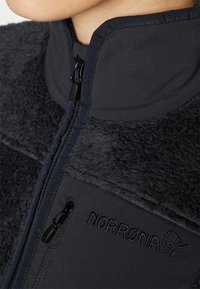 Čierna fleecová bunda s vysokým golierom, so zipsom na prednej strane a vyšitým logom "NORRØNA" na hrudi, s jemnou textúrou a kontrastnými materiálmi.
