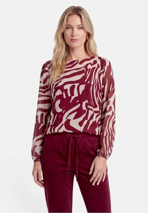 Femme aux longs cheveux blonds porte un haut à manches longues à motif abstrait bordeaux et beige et un pantalon bordeaux assorti.