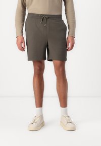 Shorts khaki à coupe décontractée, taille élastique et cordon de serrage. Associés à des baskets beige et des chaussettes blanches, le tout sur fond uni.