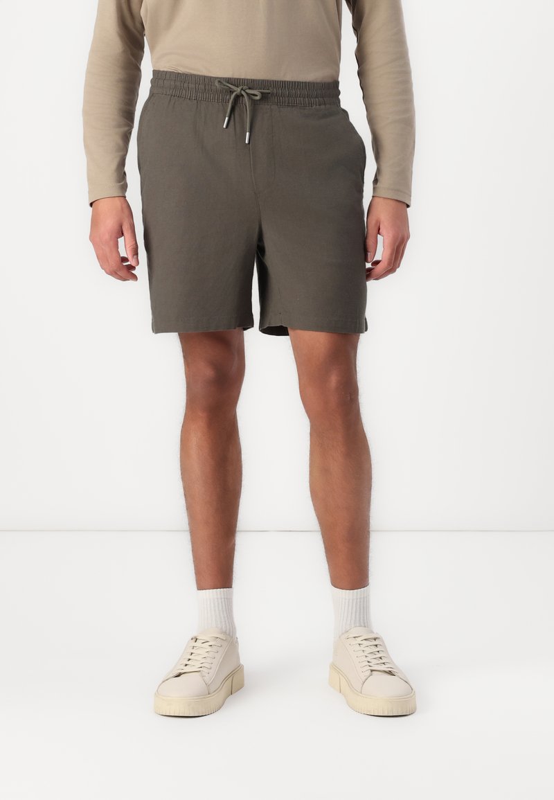 Shorts khaki à coupe décontractée, taille élastique et cordon de serrage. Associés à des baskets beige et des chaussettes blanches, le tout sur fond uni.