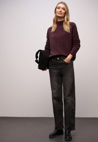 Weinroter Pullover mit hohem Kragen, kombiniert mit schwarzen Jeans mit geradem Bein. Das Modell hält eine schwarze Tasche. Glatte Texturen und neutrale Farbtöne.