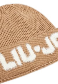 Karamelkleurige gebreide beanie met onderzijde waarop witte fuzzy tekst gedeeltelijk zichtbaar is tegen een witte achtergrond.